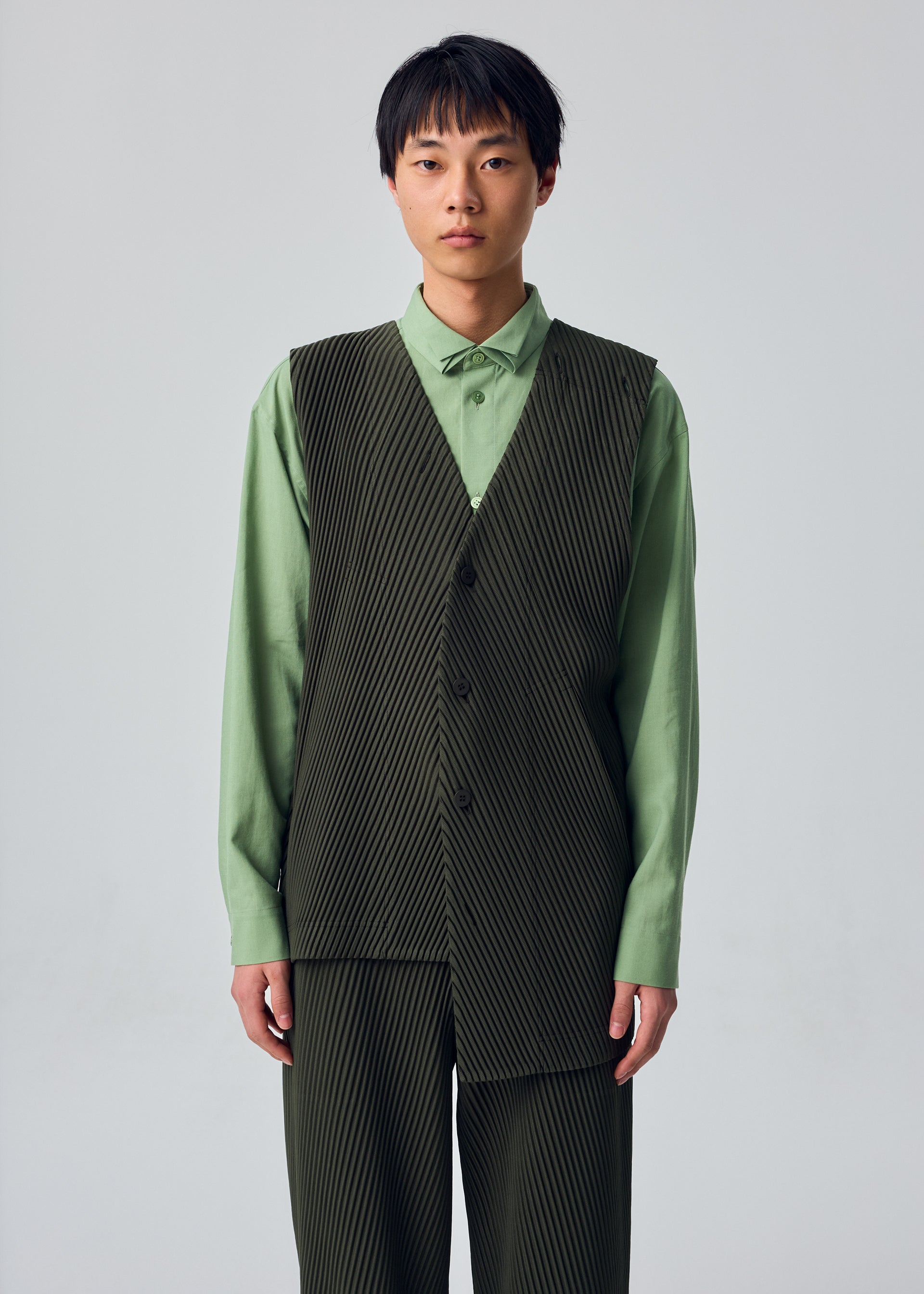 Zoomed view A model wears HOMME PLISSÉ ISSEY MIYAKE’s SLASH ensemble: a light green shirt paired with a dark green, pinstriped vest and matching pants featuring subtle diagonal pleats and slash pockets. Posed against a plain light background, the model gazes at the camera with a neutral expression.