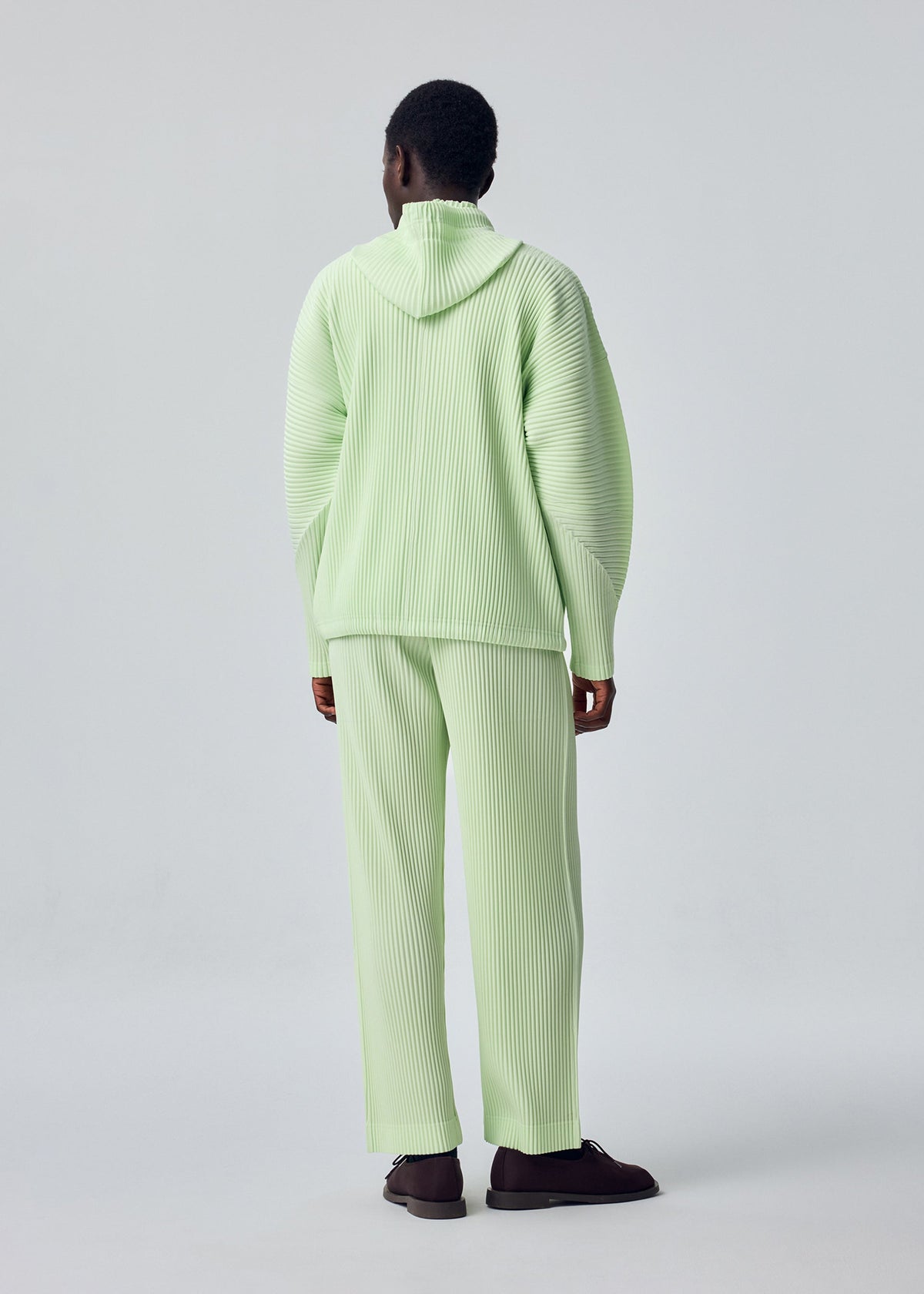 A person faces away, wearing HOMME PLISSÉ ISSEY MIYAKE’s MONTHLY COLORS : JULY light green ribbed hoodie and matching straight pants with a drawcord waist, paired with dark shoes, set against a plain light gray background. view 6 of 7