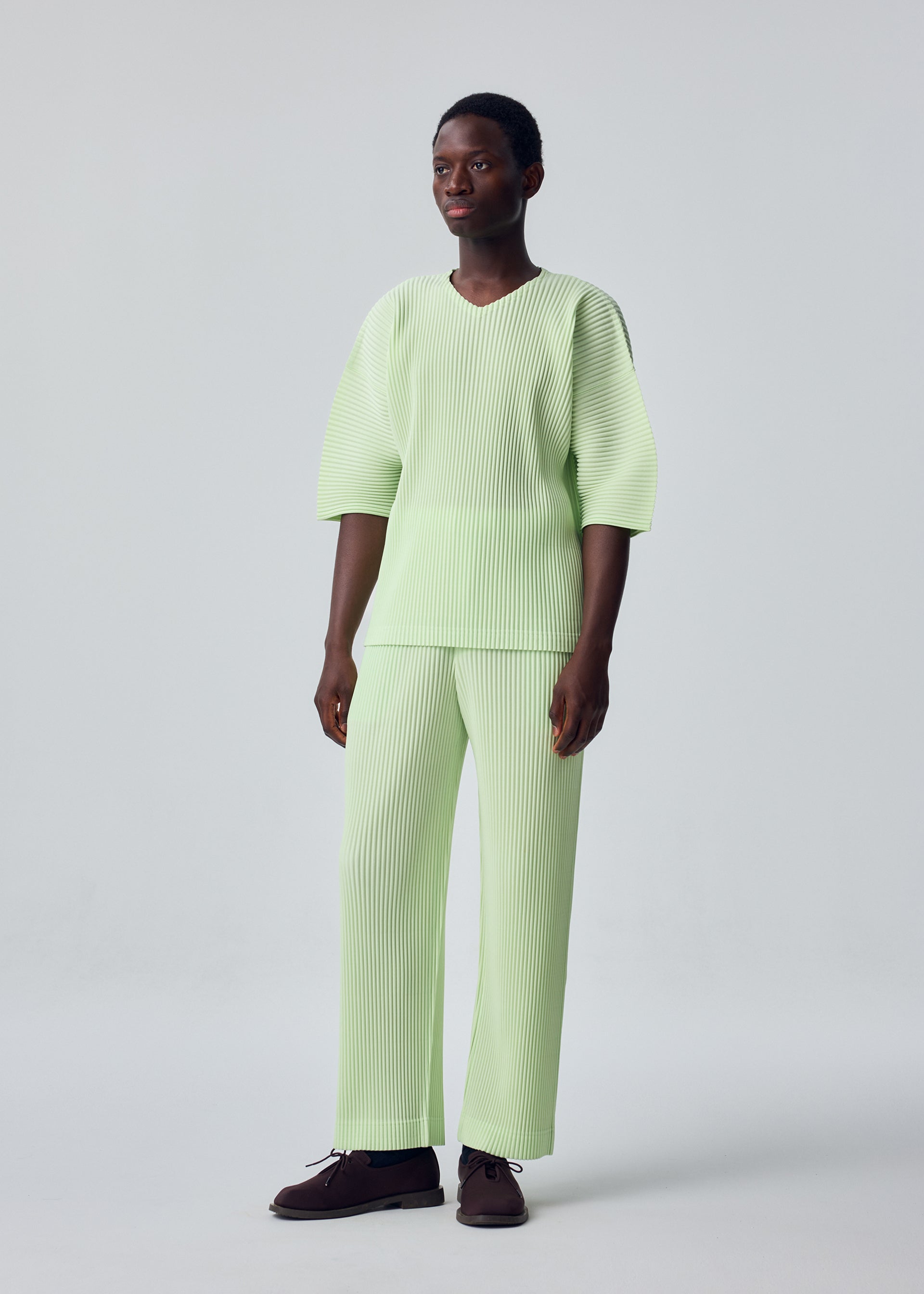 Zoomed view A person with short hair wears the HOMME PLISSÉ ISSEY MIYAKE MONTHLY COLORS: JULY set—pastel green, ribbed, loose-fitting top with elbow-length sleeves and matching straight pants in quick-drying polyester—paired with dark shoes.