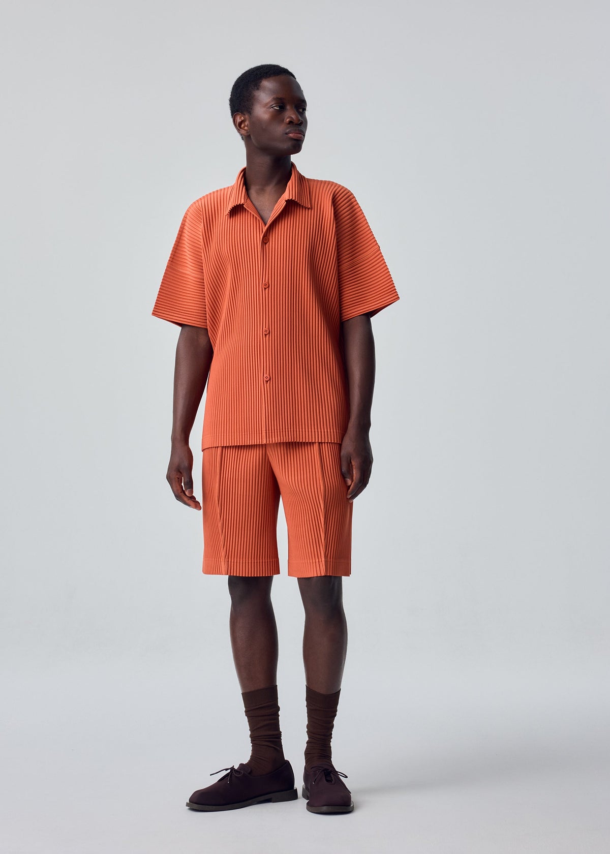 A person stands against a plain light background wearing the HOMME PLISSÉ ISSEY MIYAKE MONTHLY COLORS : AUGUST set—an orange, short-sleeved, button-up shirt and matching quick-drying polyester shorts with vertical stripes—paired with brown socks and dark brown shoes. view 6 of 7