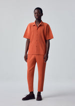Wearing the HOMME PLISSÉ ISSEY MIYAKE MONTHLY COLORS : AUGUST set, a person stands against a plain background in an orange short-sleeve button-up shirt and matching orange tapered pants with subtle vertical stripes, paired with dark brown shoes for lightweight comfort.