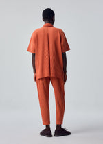A person faces away, dressed in the HOMME PLISSÉ ISSEY MIYAKE MONTHLY COLORS : AUGUST collection, featuring an oversized orange short-sleeve shirt and matching quick-drying orange tapered pants with vertical stripes. The background is plain and light-colored.