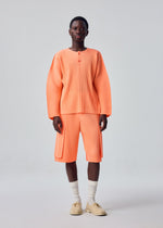 A person stands facing forward against a plain light gray background, dressed in the HOMME PLISSÉ ISSEY MIYAKE MONTHLY COLORS : SEPTEMBER set—a loose, peach-colored ribbed long-sleeve top paired with matching knee-length polyester shorts—styled with white socks and beige shoes.