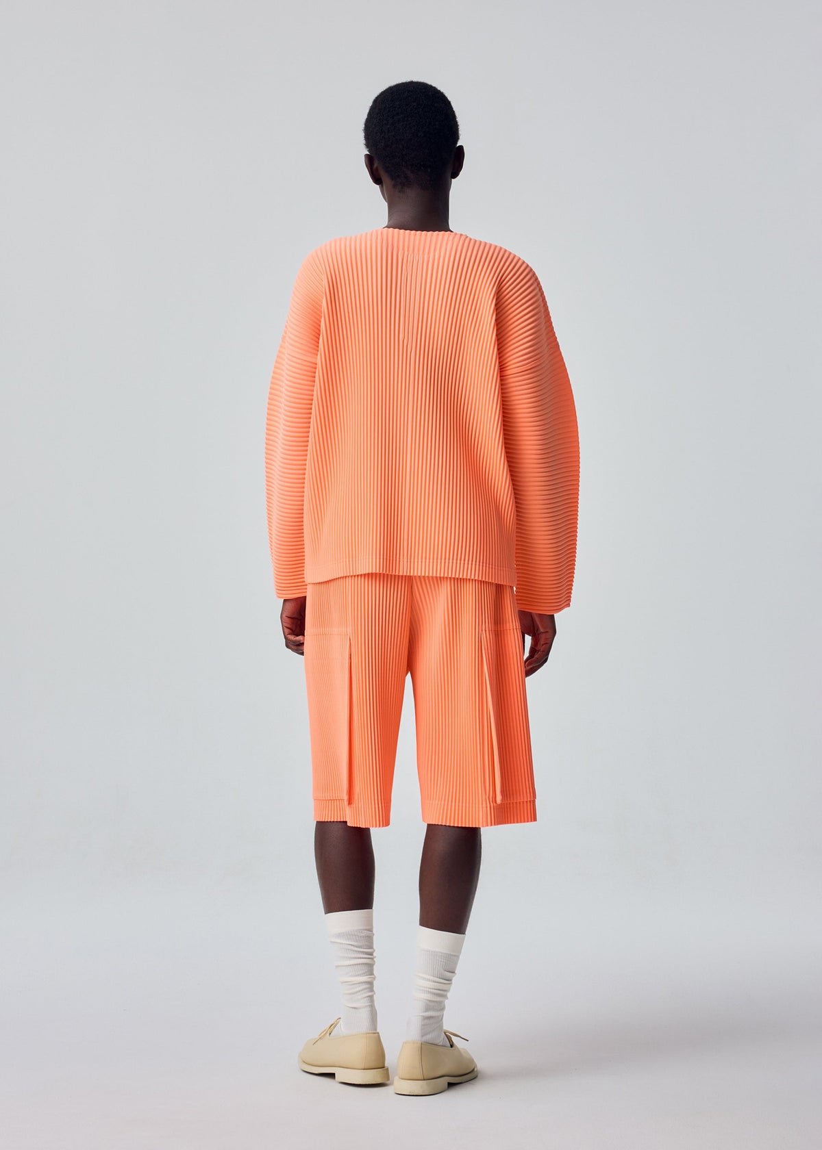 A person stands with their back to the camera, wearing HOMME PLISSÉ ISSEY MIYAKE’s MONTHLY COLORS : SEPTEMBER oversized pleated peach top with matching long polyester shorts, paired with white socks and beige shoes against a plain light gray background. view 7 of 7