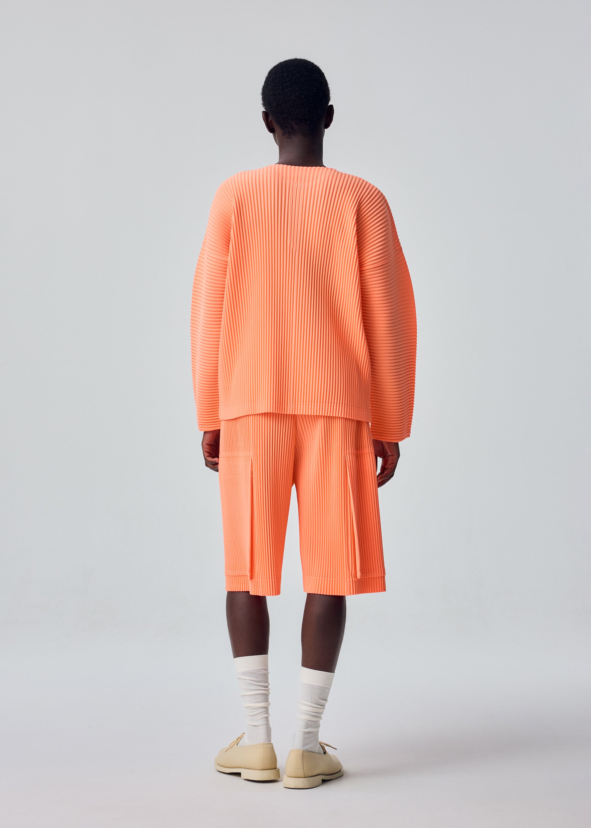Zoomed view A person stands with their back to the camera, wearing HOMME PLISSÉ ISSEY MIYAKE’s MONTHLY COLORS : SEPTEMBER oversized pleated peach top with matching long polyester shorts, paired with white socks and beige shoes against a plain light gray background.