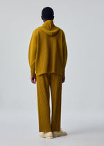 A person stands facing away, dressed in HOMME PLISSÉ ISSEY MIYAKE MONTHLY COLORS : OCTOBER mustard yellow pleated hoodie and matching quick-drying polyester straight pants, with cream shoes against a light gray background.