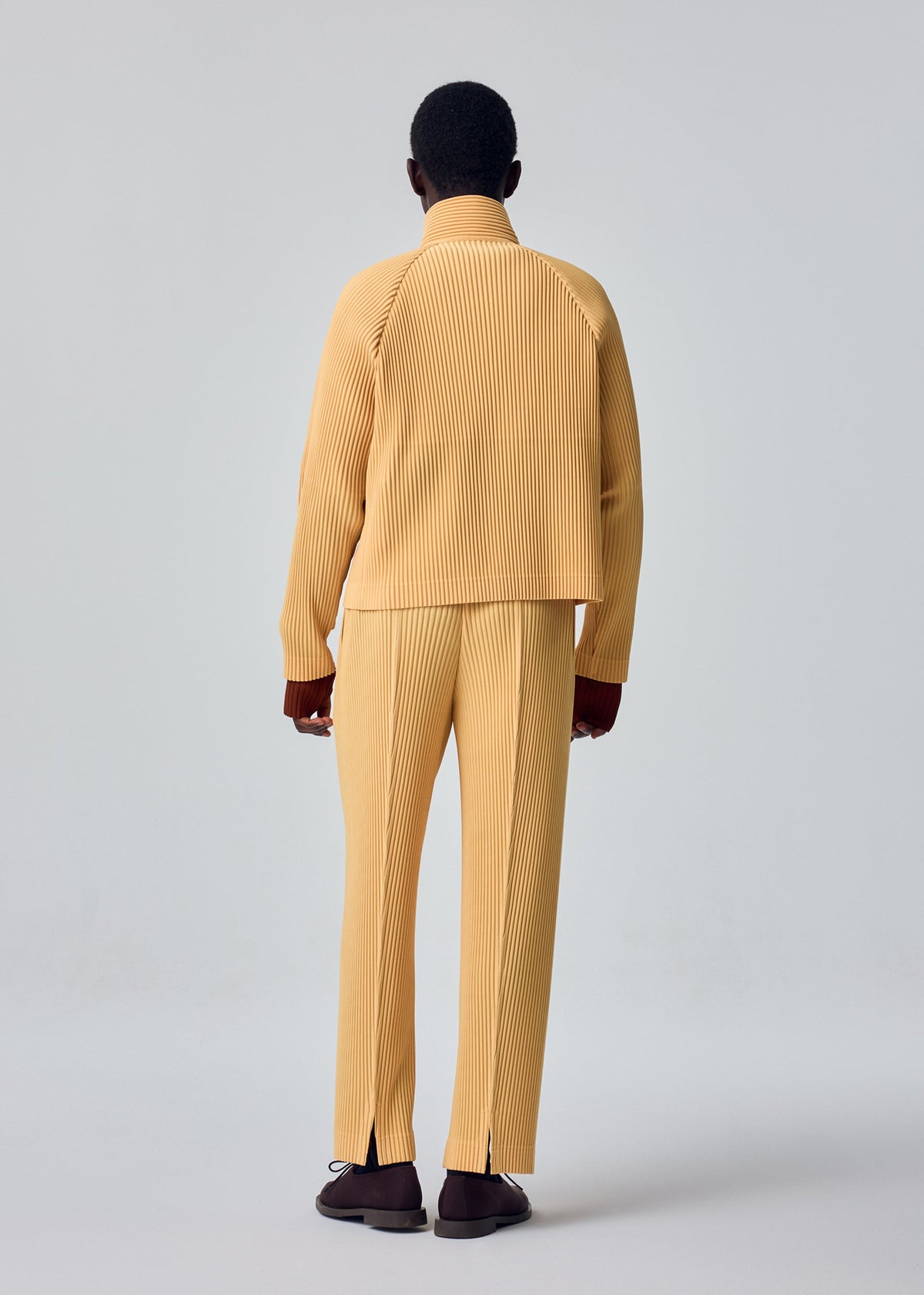 A person stands facing away, wearing the HOMME PLISSÉ ISSEY MIYAKE TAILORED PLEATS 1 set in yellow—comprising a ribbed jacket and center-pressed pants—with brown shoes, against a plain light gray background. view 6 of 7