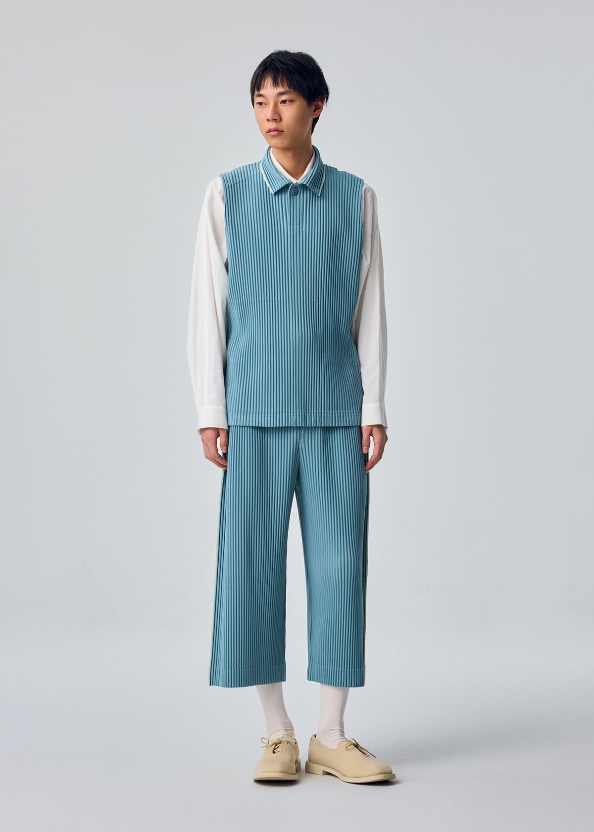 A person stands against a plain background wearing the EMBROIDERY STRIPES set from HOMME PLISSÉ ISSEY MIYAKE—a blue pinstriped sleeveless vest and matching embroidered cropped pants—layered over a white shirt, with white socks and beige shoes. view 5 of 7