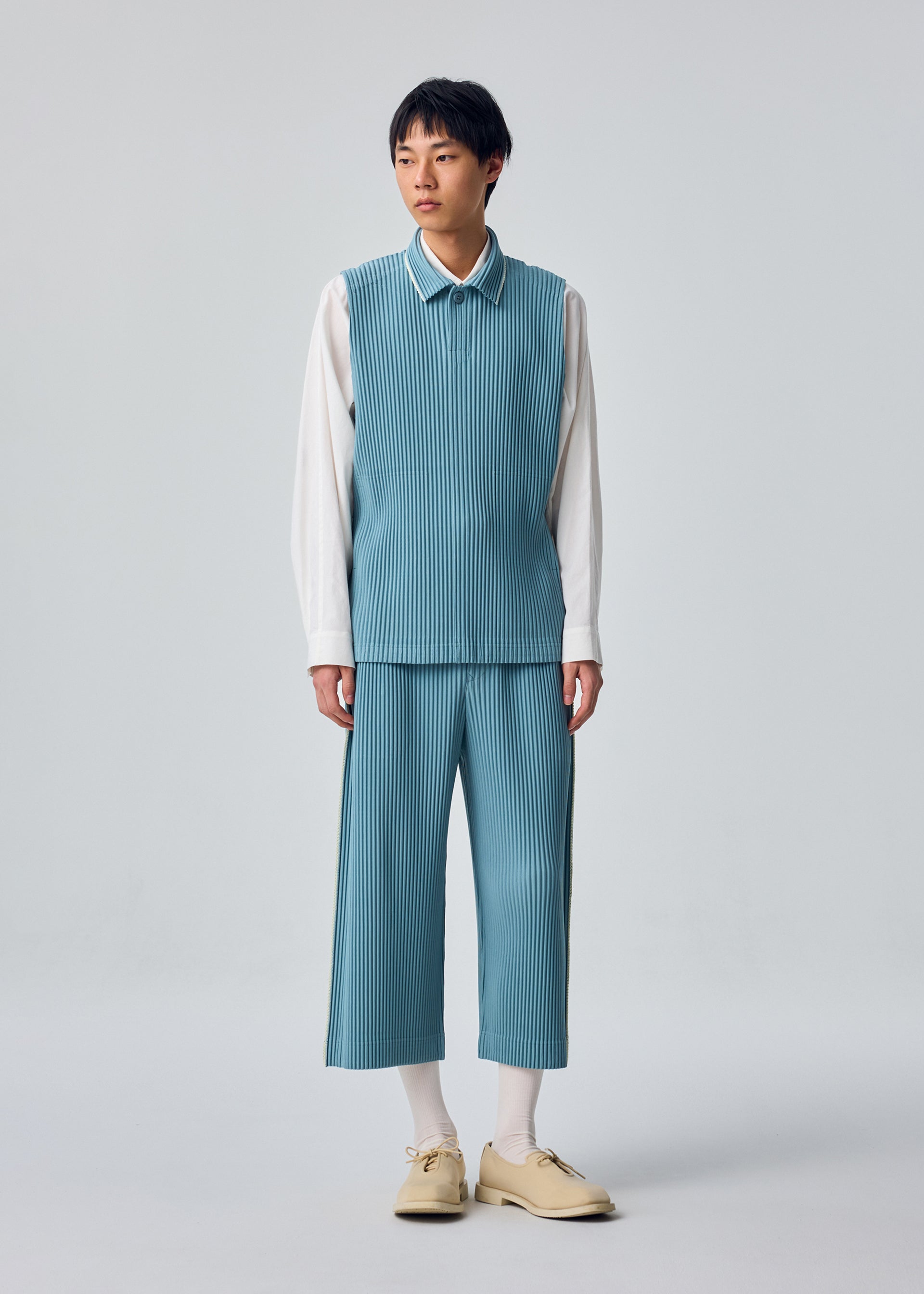 Zoomed view A person stands against a plain background wearing the EMBROIDERY STRIPES set from HOMME PLISSÉ ISSEY MIYAKE—a blue pinstriped sleeveless vest and matching embroidered cropped pants—layered over a white shirt, with white socks and beige shoes.