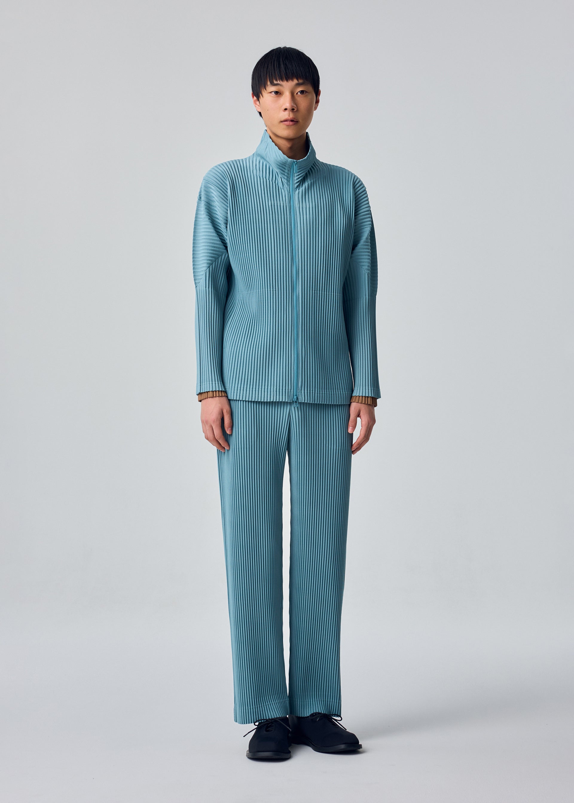 Zoomed view A person stands against a plain background wearing the COLOR PLEATS 2 set from HOMME PLISSÉ ISSEY MIYAKE—a matching light blue pleated jacket and sporty straight pants crafted from quick-drying polyester—paired with black shoes, looking straight ahead with a neutral expression.