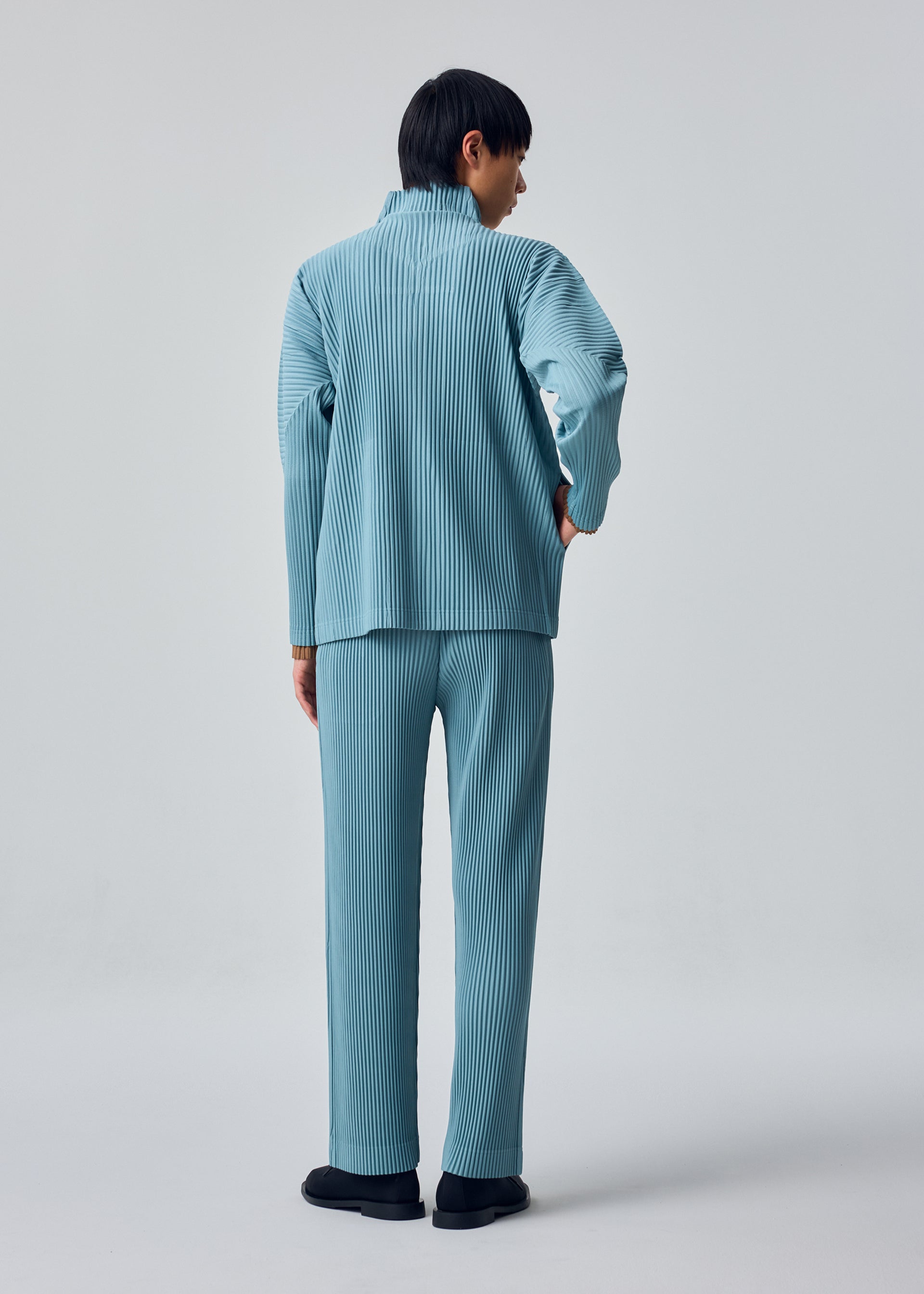 Zoomed view A person stands facing away, wearing the HOMME PLISSÉ ISSEY MIYAKE COLOR PLEATS 2 set: a light blue, pleated matching top and sporty straight pants with a drawcord waist crafted from quick-drying polyester, paired with black shoes, against a plain light gray background.