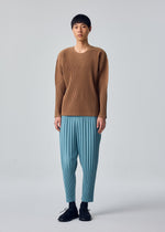 A person stands against a plain background, wearing the COLOR PLEATS 2 ribbed tan sweater with long sleeves from HOMME PLISSÉ ISSEY MIYAKE and loose, sporty light blue pants featuring a drawcord waist. They have short black hair, are wearing black shoes, and have a neutral facial expression.