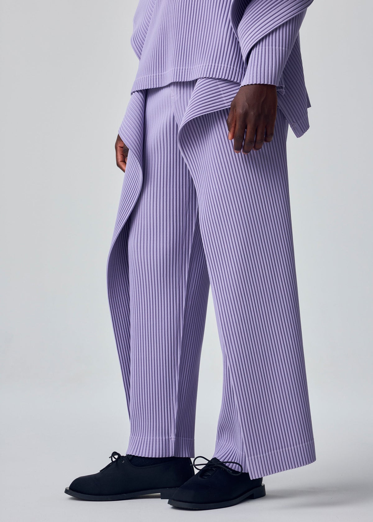 A person showcases the HOMME PLISSÉ ISSEY MIYAKE MOTION BLUR set—loose, pleated jersey pants with a drawcord waist and matching lavender top—paired with black shoes, against a light gray background, highlighting its experimental silhouette. view 7 of 8