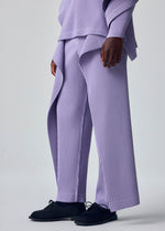 A person showcases the HOMME PLISSÉ ISSEY MIYAKE MOTION BLUR set—loose, pleated jersey pants with a drawcord waist and matching lavender top—paired with black shoes, against a light gray background, highlighting its experimental silhouette.