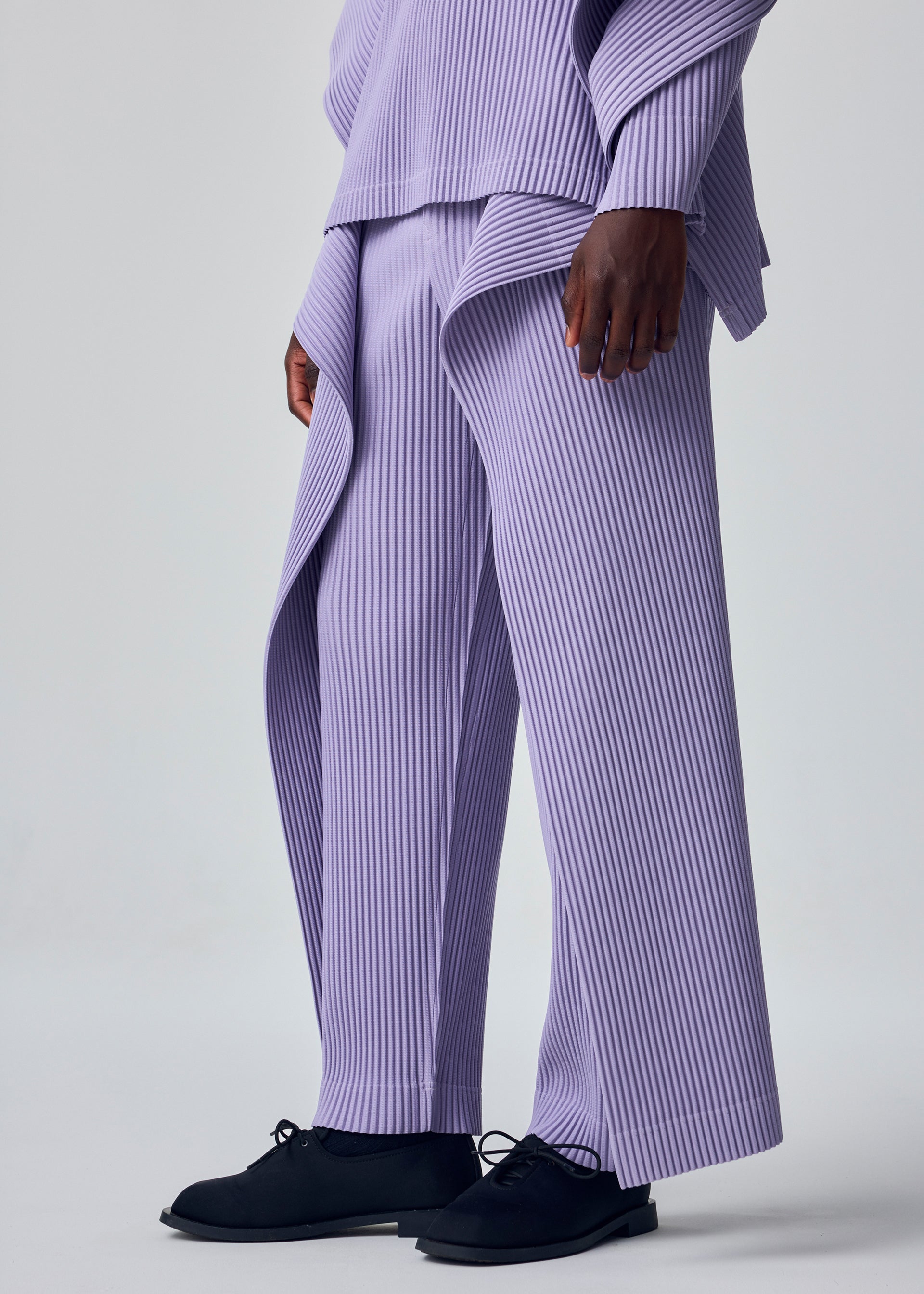 Zoomed view A person showcases the HOMME PLISSÉ ISSEY MIYAKE MOTION BLUR set—loose, pleated jersey pants with a drawcord waist and matching lavender top—paired with black shoes, against a light gray background, highlighting its experimental silhouette.