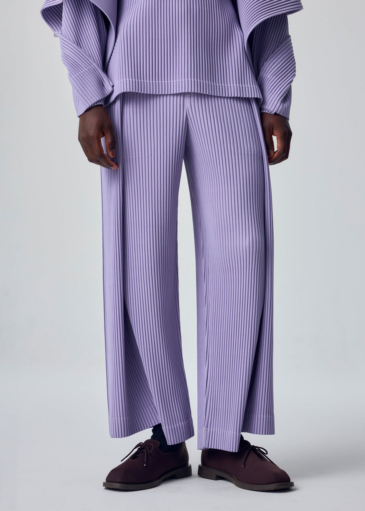A person wears the HOMME PLISSÉ ISSEY MIYAKE MOTION BLUR set—lavender rib-knit pleated jersey pants and a long-sleeve top—with dark brown shoes. The image is shoulder-cropped against a plain background. view 8 of 8