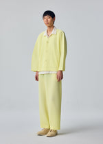 A person stands in a studio against a plain light gray background, wearing the SLASH suit by HOMME PLISSÉ ISSEY MIYAKE—a loose-fit, light yellow ensemble featuring diagonal pleats and wide pants with a drawcord waist—paired with beige shoes.