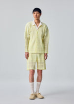 A person stands against a plain background, wearing the HOMME PLISSÉ ISSEY MIYAKE LACE GRID PLEATS—a pale yellow textured jacket with matching cotton-blend polyester shorts featuring a drawcord waist—styled with a white shirt, cream socks, and beige shoes.