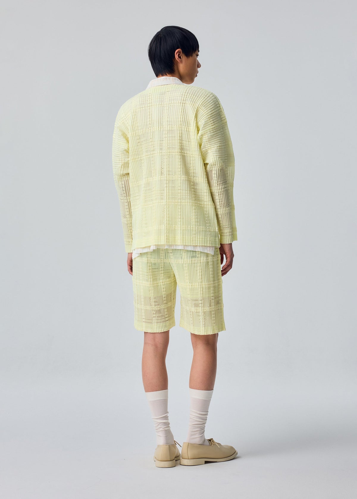 A person with short dark hair stands facing away, wearing the pale yellow LACE GRID PLEATS jacket and matching lace shorts by HOMME PLISSÉ ISSEY MIYAKE, paired with long white socks and beige shoes, against a plain light background. view 6 of 7