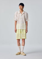 A person stands against a plain background wearing the HOMME PLISSÉ ISSEY MIYAKE LACE GRID PLEATS short-sleeve button-up shirt paired with matching yellow lace shorts featuring a drawcord waist, complemented by long white socks and beige shoes.