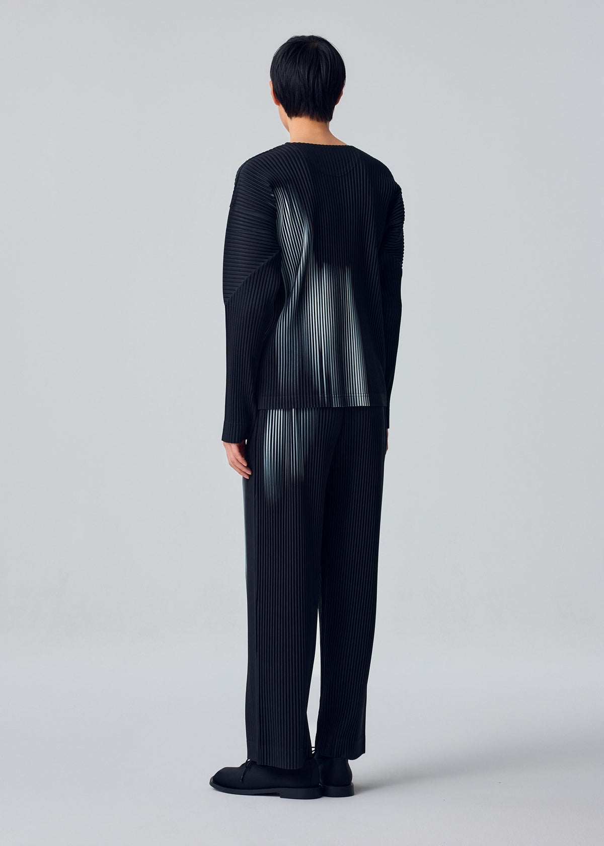 Person stands with their back to the camera, wearing HOMME PLISSÉ ISSEY MIYAKE IN THE SHADOW—a loose-fitting black pleated long-sleeve top and straight pants—against a plain light gray background. view 5 of 5