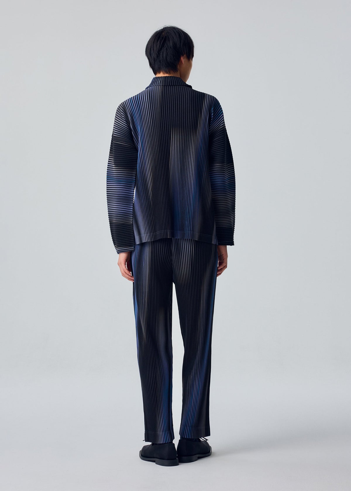 A person faces away from the camera, wearing the HOMME PLISSÉ ISSEY MIYAKE BLEND OF LIGHTS set—loose long-sleeved top and striped drawcord pants with black shoes—against a plain light gray background. view 4 of 5