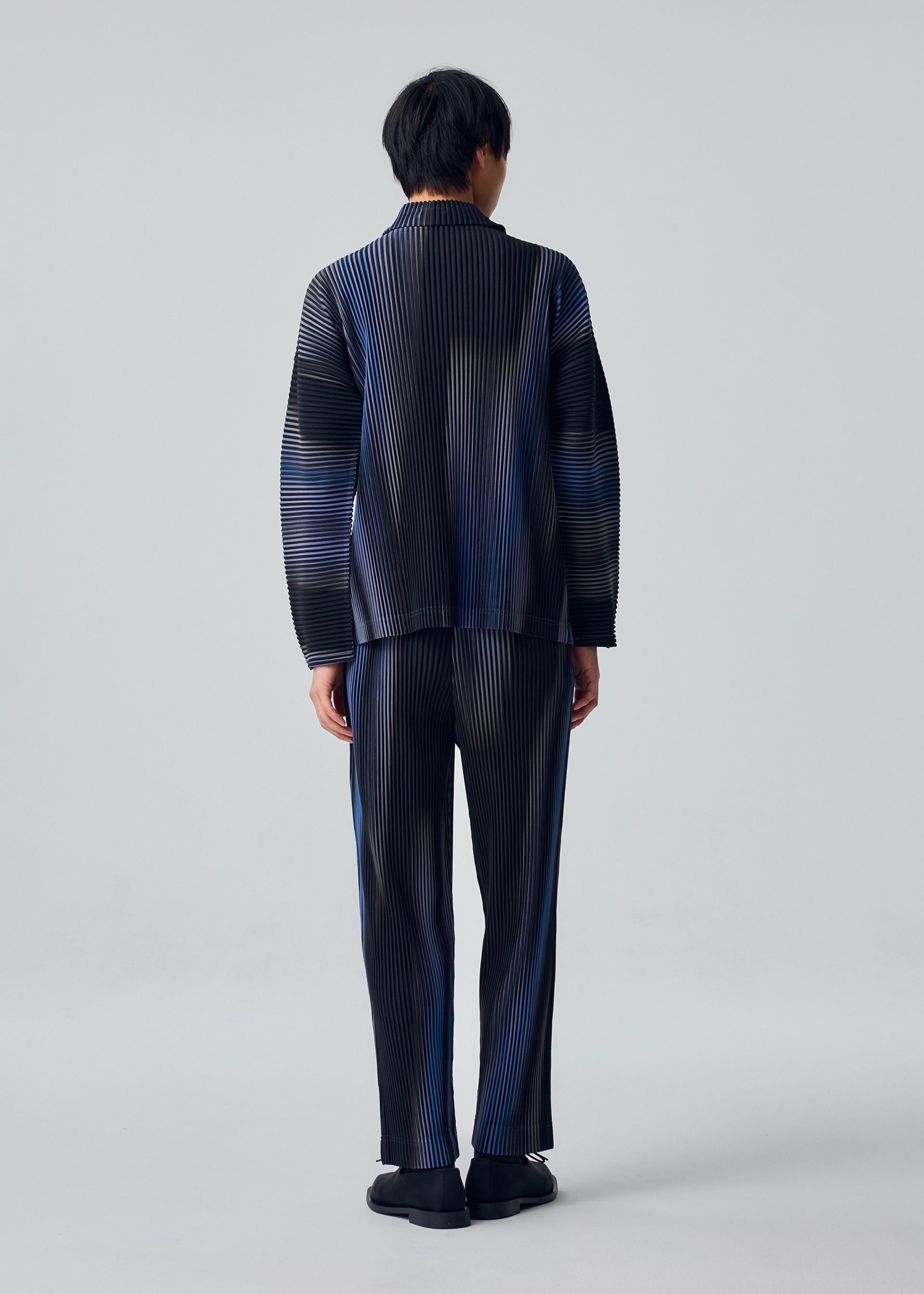Zoomed view A person faces away from the camera, wearing the HOMME PLISSÉ ISSEY MIYAKE BLEND OF LIGHTS set—loose long-sleeved top and striped drawcord pants with black shoes—against a plain light gray background.
