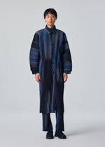 A person stands against a plain background wearing the BLEND OF LIGHTS coat and matching straight pants with vertical black and blue stripes by HOMME PLISSÉ ISSEY MIYAKE, styled with dark shoes for a modern, minimalist look.