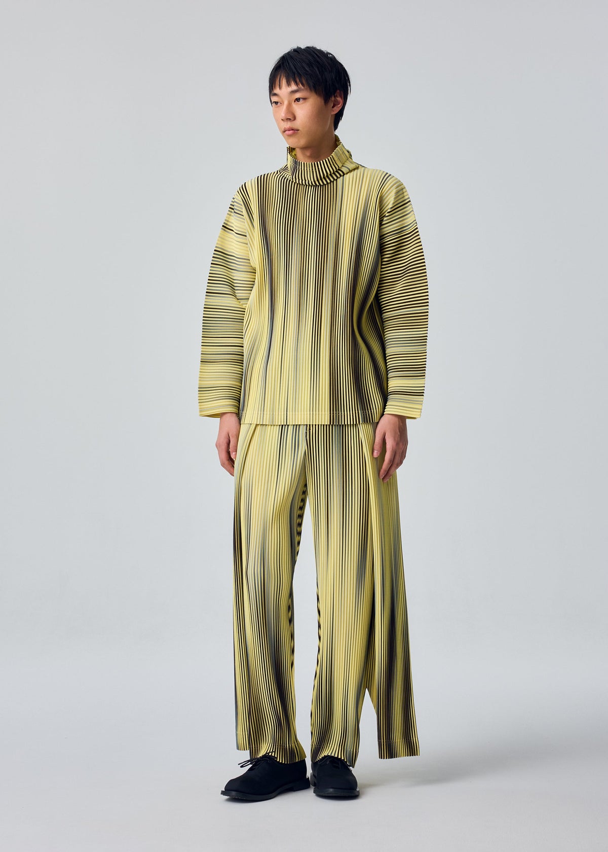 A person stands against a plain background wearing the HOMME PLISSÉ ISSEY MIYAKE "BLEND OF LIGHTS" set: a matching yellow and black vertically striped top and wide-legged pants with a drawcord waist, paired with black shoes. view 6 of 8