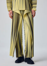 A person wears the HOMME PLISSÉ ISSEY MIYAKE BLEND OF LIGHTS set—yellow and black vertical striped pants with front slits and a drawcord waist, paired with the matching long-sleeve top—creating a geometric, flowing effect. Black shoes complete the look.