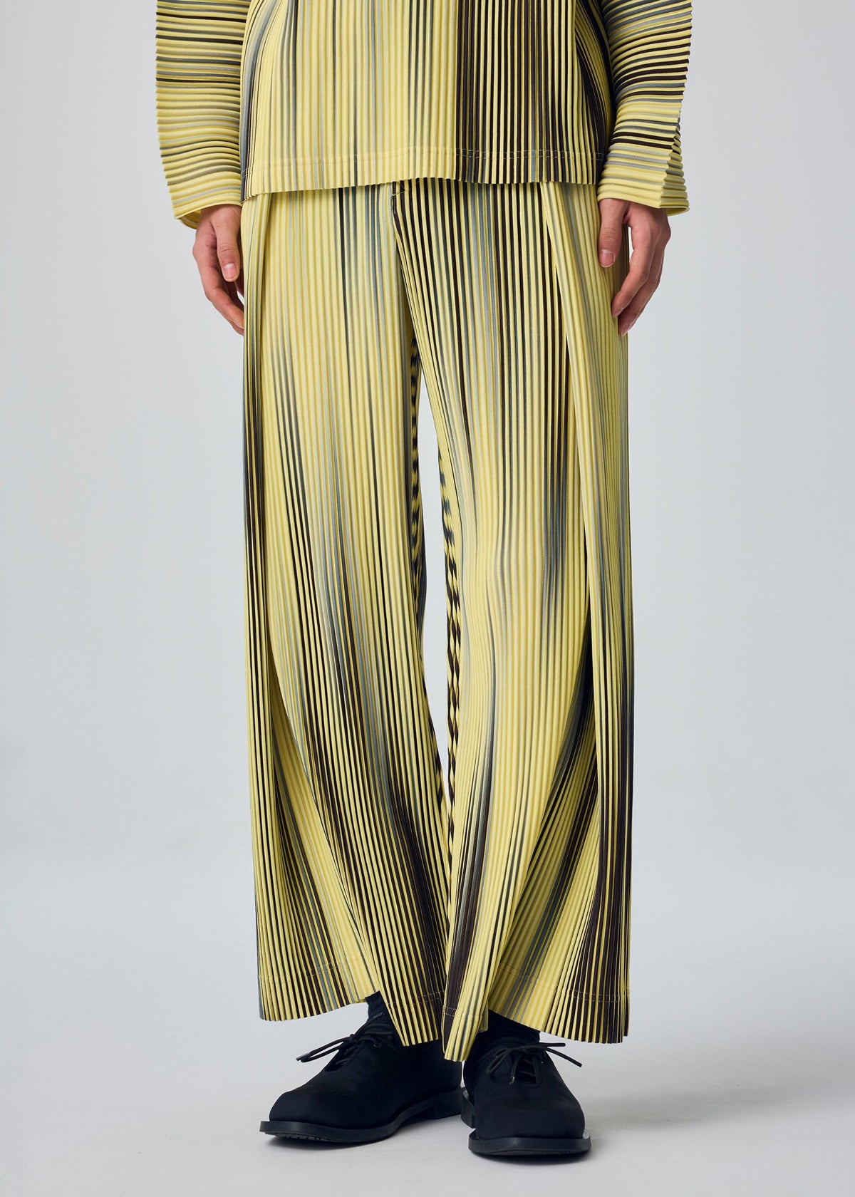 A person wears HOMME PLISSÉ ISSEY MIYAKE's BLEND OF LIGHTS yellow pants with black vertical stripes, pleats, and drawcord waist, a matching top partially visible, and black shoes against a plain light background. view 8 of 8
