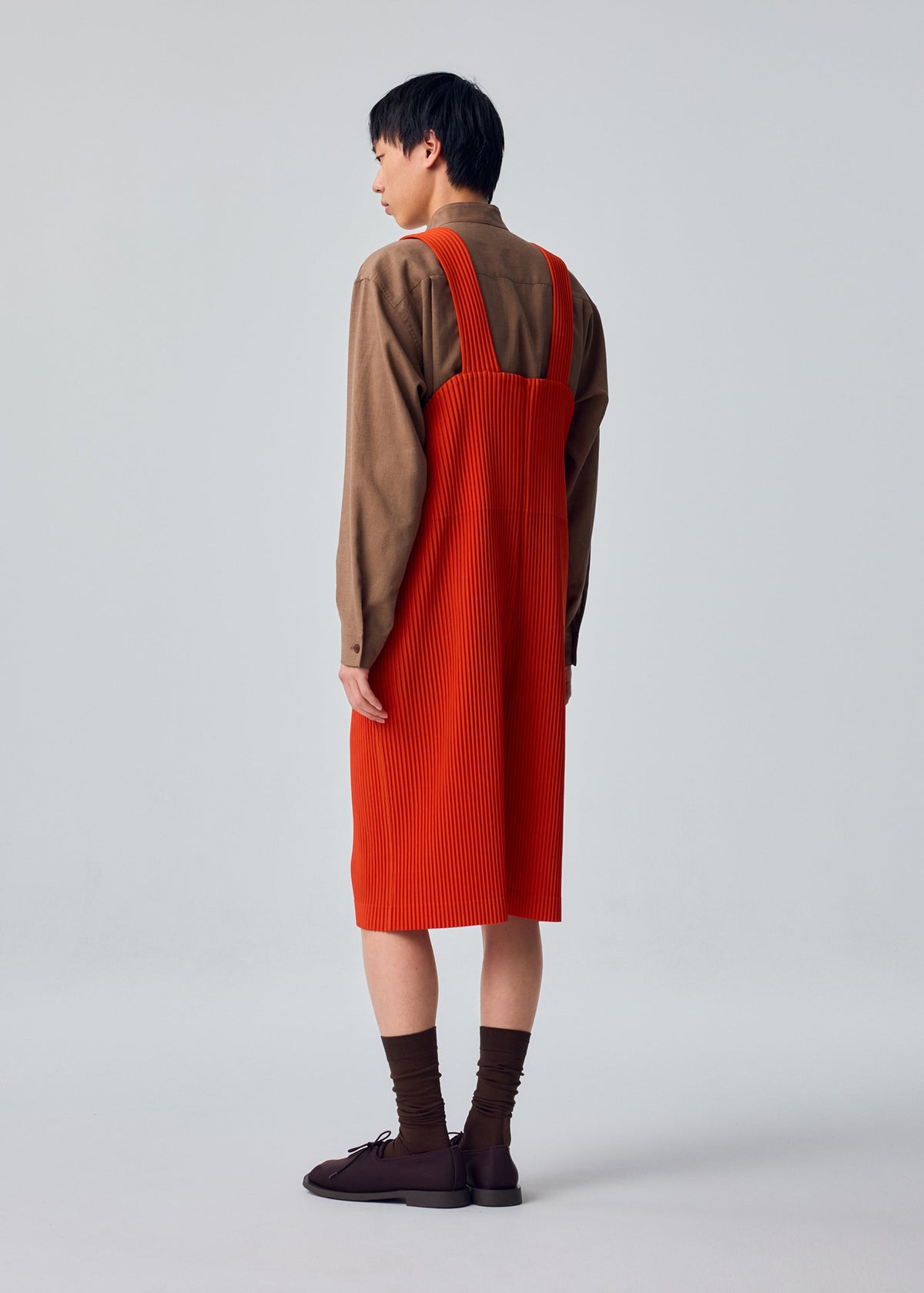 A person faces away, wearing HOMME PLISSÉ ISSEY MIYAKE’s PLEATS BOTTOMS 1, styled with a long-sleeved brown shirt, bright orange knee-length pinafore dress with pockets, brown socks, and dark shoes against a light background. view 5 of 5