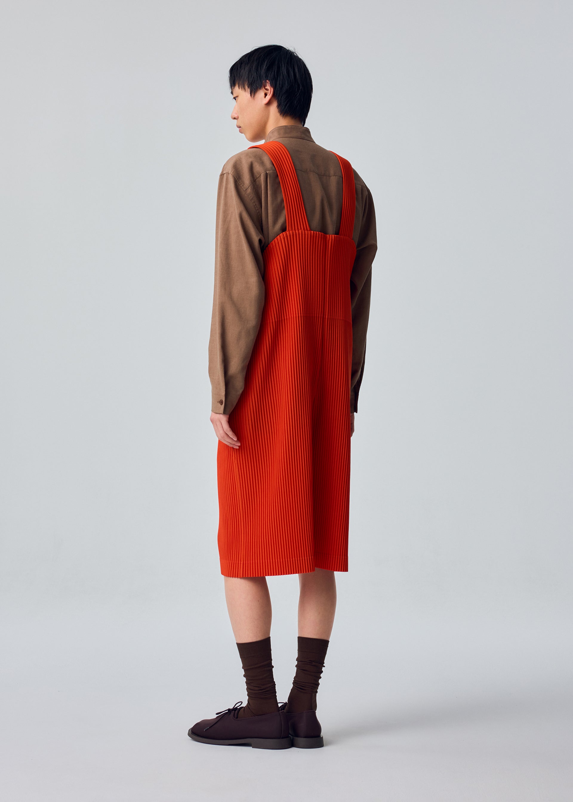 Zoomed view A person faces away, wearing HOMME PLISSÉ ISSEY MIYAKE’s PLEATS BOTTOMS 1, styled with a long-sleeved brown shirt, bright orange knee-length pinafore dress with pockets, brown socks, and dark shoes against a light background.