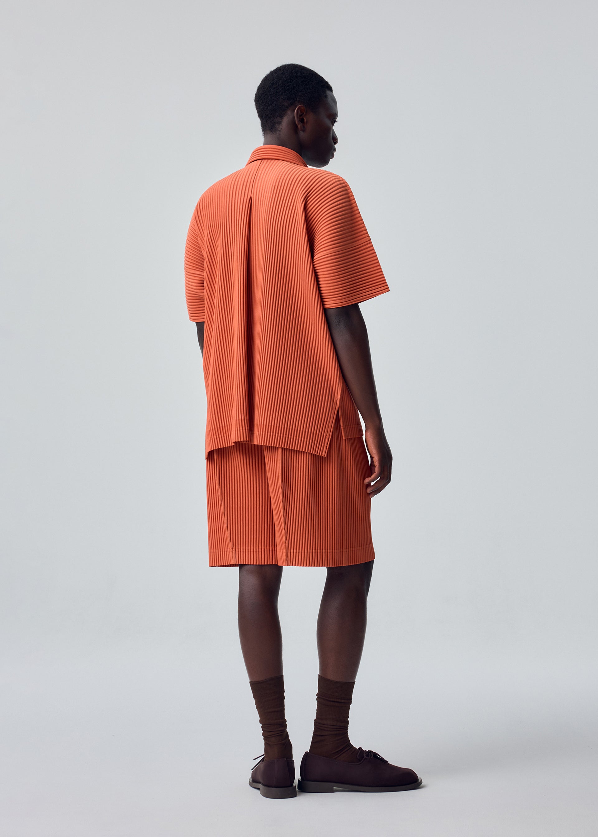 Zoomed view A person stands facing away, wearing the HOMME PLISSÉ ISSEY MIYAKE MONTHLY COLORS : AUGUST set—an orange, short-sleeved, pleated open collar shirt with matching shorts crafted from quick-drying polyester for lightweight comfort—paired with brown socks and sandals. The background is plain light gray.
