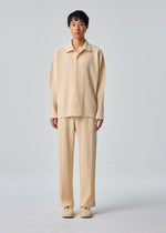 A person stands facing forward in the MONTHLY COLORS : OCTOBER set by HOMME PLISSÉ ISSEY MIYAKE—beige, pleated open collar shirt and matching pants with beige shoes, against a light gray background.