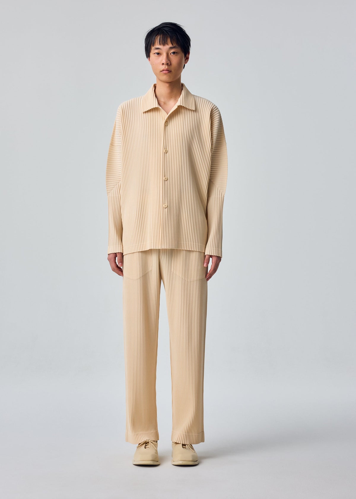 Zoomed view A person stands facing forward in the MONTHLY COLORS : OCTOBER set by HOMME PLISSÉ ISSEY MIYAKE—beige, pleated open collar shirt and matching pants with beige shoes, against a light gray background.