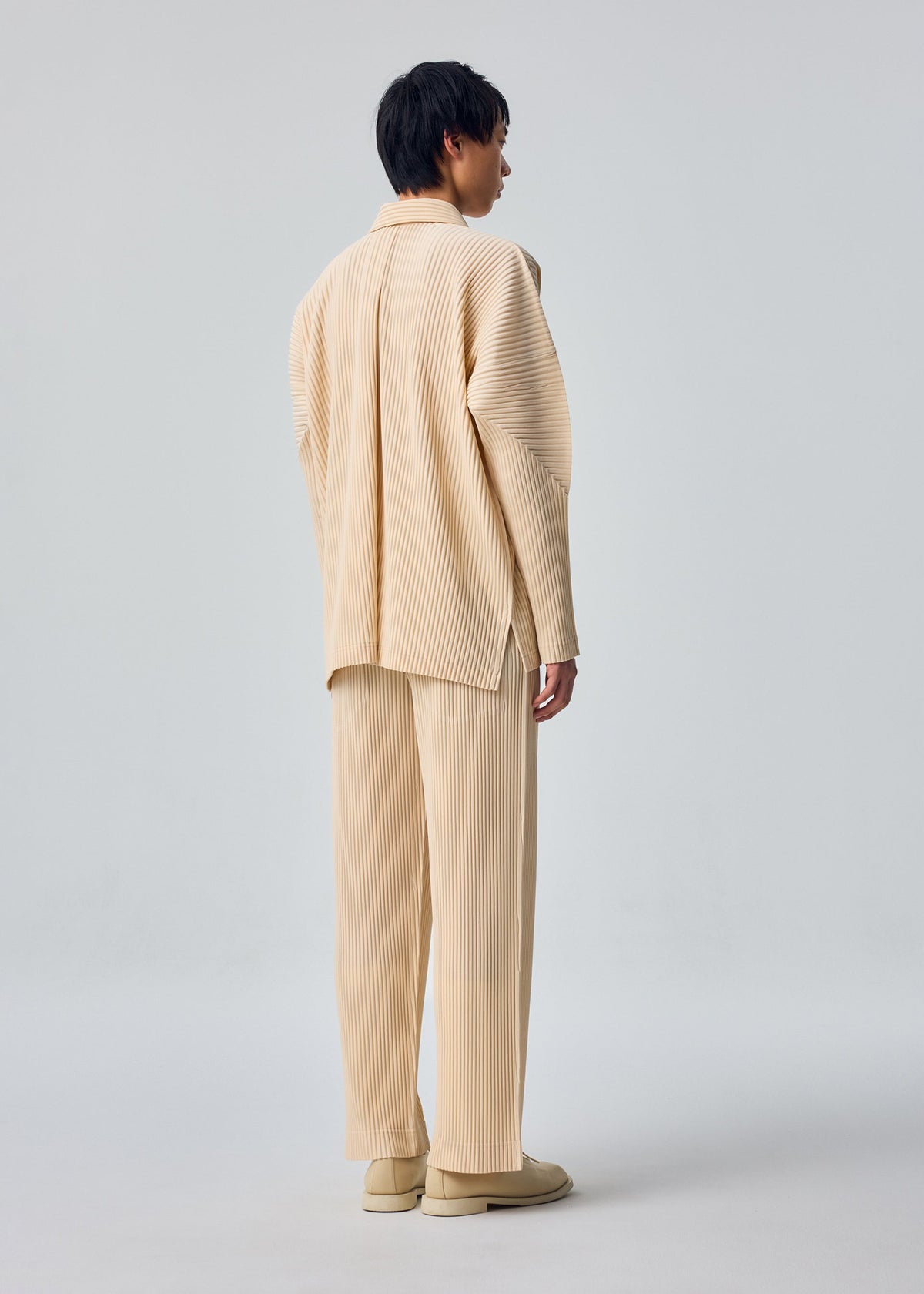A model faces away, dressed in HOMME PLISSÉ ISSEY MIYAKE MONTHLY COLORS : OCTOBER—a loose beige ribbed suit with matching pants and shoes, open collar shirt, set against a plain light gray background. view 7 of 7