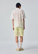 A person stands facing away, wearing the LACE GRID PLEATS shorts by HOMME PLISSÉ ISSEY MIYAKE—a light yellow textured design—paired with a sheer white open-collar cotton-blend polyester shirt, white socks, and beige shoes against a plain light background.