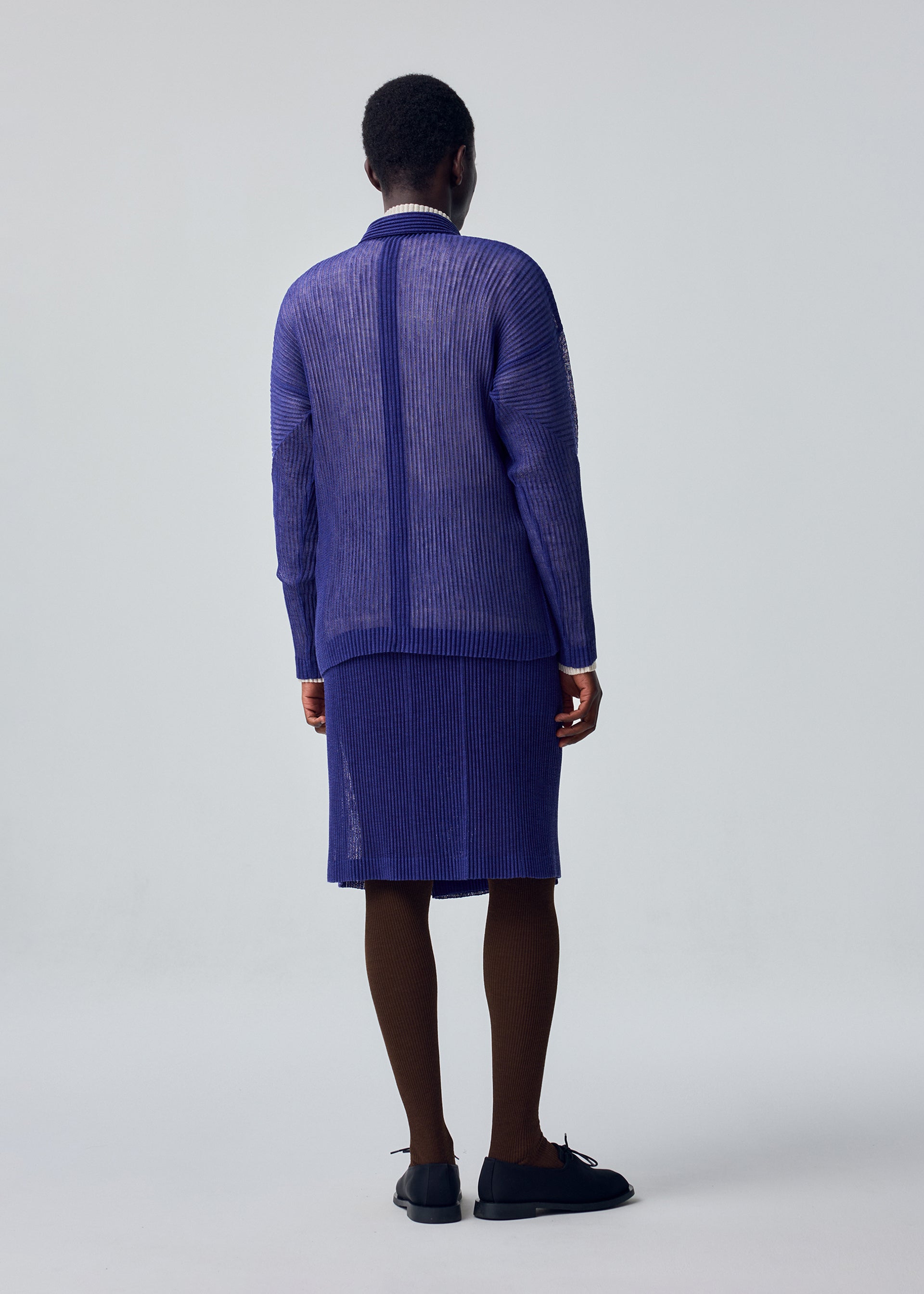 Zoomed view A person stands facing away, wearing the RUSTIC MESH sheer blue top and matching knee-length skirt by HOMME PLISSÉ ISSEY MIYAKE, layered over brown tights with black shoes, against a plain light gray background.