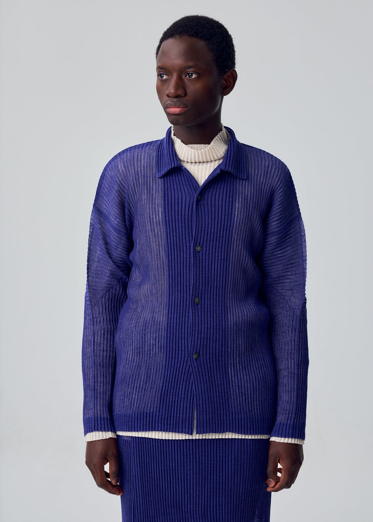A person wears the HOMME PLISSÉ ISSEY MIYAKE RUSTIC MESH long-sleeved blue button-up shirt over a cream turtleneck, standing against a plain light gray background and looking slightly to the side. view 7 of 7