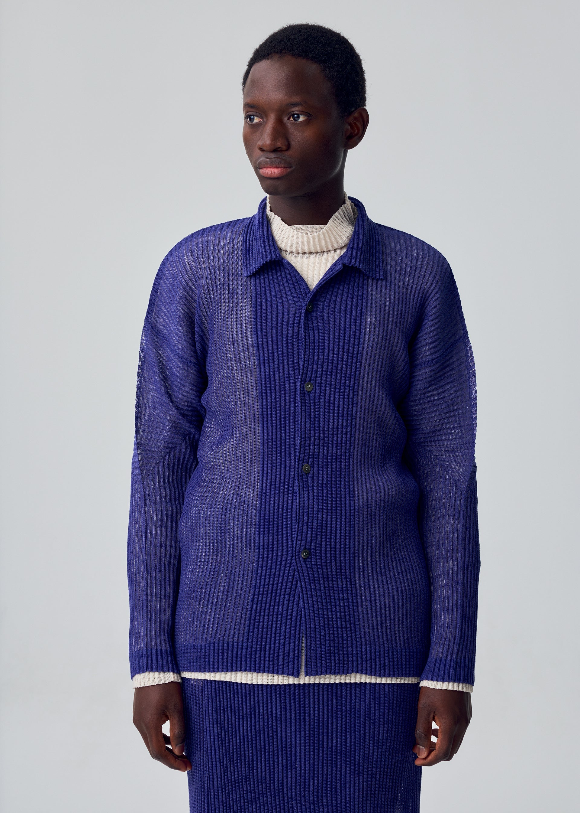 Zoomed view A person wears the HOMME PLISSÉ ISSEY MIYAKE RUSTIC MESH long-sleeved blue button-up shirt over a cream turtleneck, standing against a plain light gray background and looking slightly to the side.