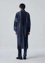 A person stands facing away, wearing HOMME PLISSÉ ISSEY MIYAKE’s BLEND OF LIGHTS—a long, pleated coat and matching striped pants in black, blue, and gray, with black shoes. The background is plain light gray.