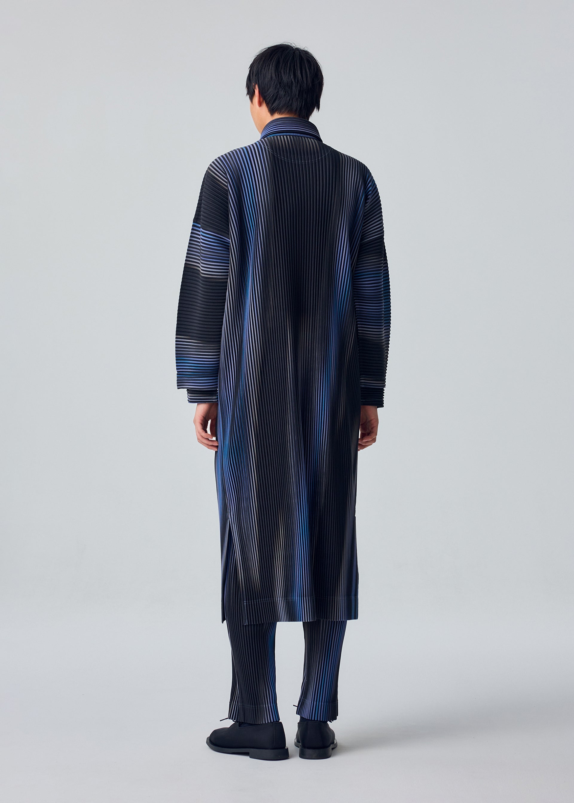 Zoomed view A person stands facing away, wearing HOMME PLISSÉ ISSEY MIYAKE’s BLEND OF LIGHTS—a long, pleated coat and matching striped pants in black, blue, and gray, with black shoes. The background is plain light gray.