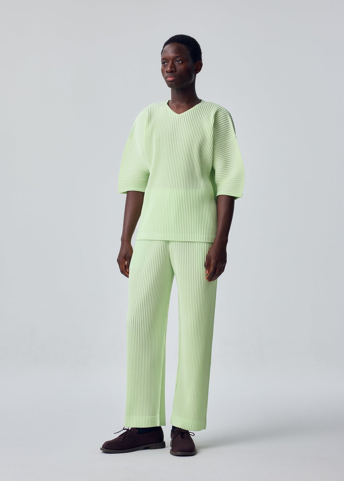 A person stands against a plain light background, wearing the HOMME PLISSÉ ISSEY MIYAKE MONTHLY COLORS: JULY set—light green, ribbed V-neck top with three-quarter sleeves and loose-fitting pants—paired with dark shoes. view 4 of 4