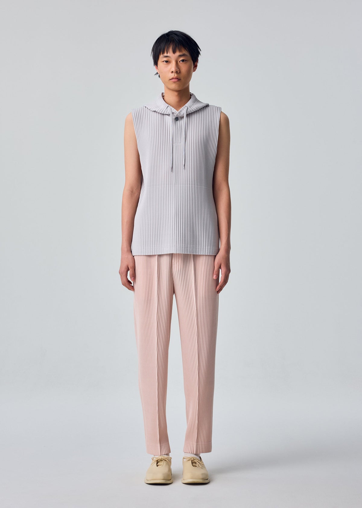 A person stands facing forward against a plain light gray background, wearing the HOMME PLISSÉ ISSEY MIYAKE MONTHLY COLORS : SEPTEMBER sleeveless hoodie in light gray—a lightweight, quick-drying polyester style—paired with loose, pale pink pleated pants and cream-colored shoes. view 6 of 7