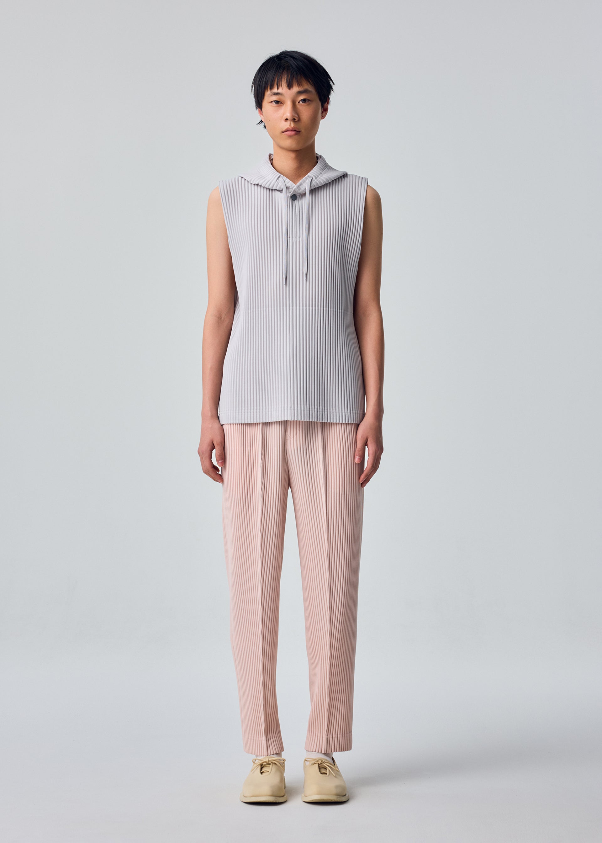 Zoomed view A person stands facing forward against a plain light gray background, wearing the HOMME PLISSÉ ISSEY MIYAKE MONTHLY COLORS : SEPTEMBER sleeveless hoodie in light gray—a lightweight, quick-drying polyester style—paired with loose, pale pink pleated pants and cream-colored shoes.