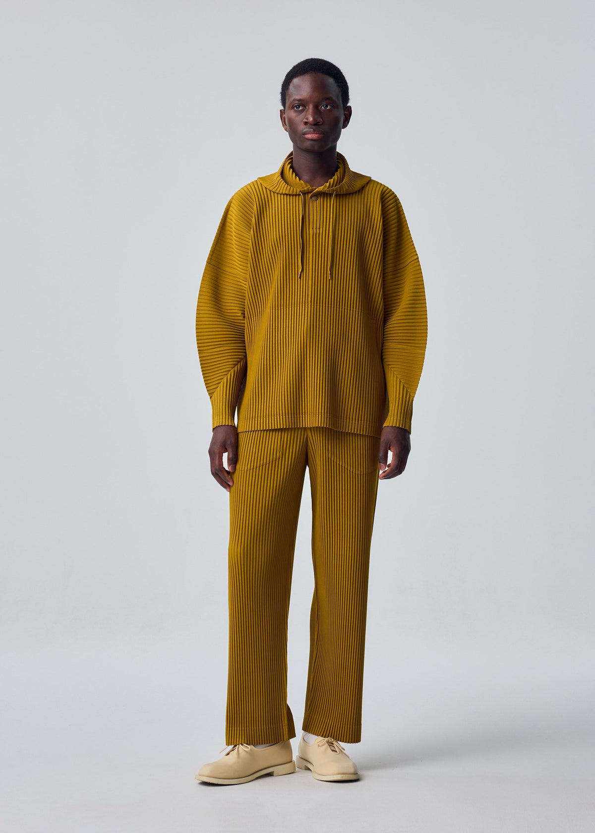 Zoomed view A person stands facing forward in the MONTHLY COLORS : OCTOBER set by HOMME PLISSÉ ISSEY MIYAKE—mustard yellow, pleated long-sleeve top with collar and matching loose pants. Light beige shoes, neutral expression, plain light gray backdrop.