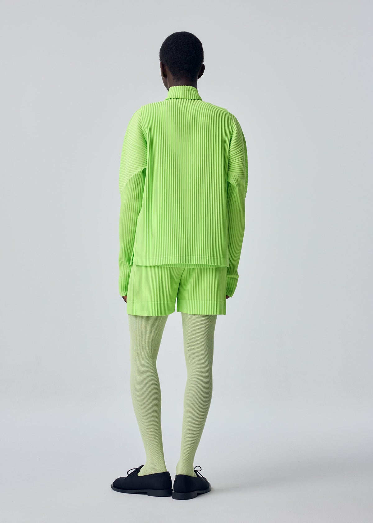 A person stands with their back to the camera against a plain light background, dressed in the HOMME PLISSÉ ISSEY MIYAKE COLOR PLEATS 1 set: a pleated neon green jacket with thumbhole sleeves and matching shorts, paired with pale green tights and black shoes. view 8 of 9