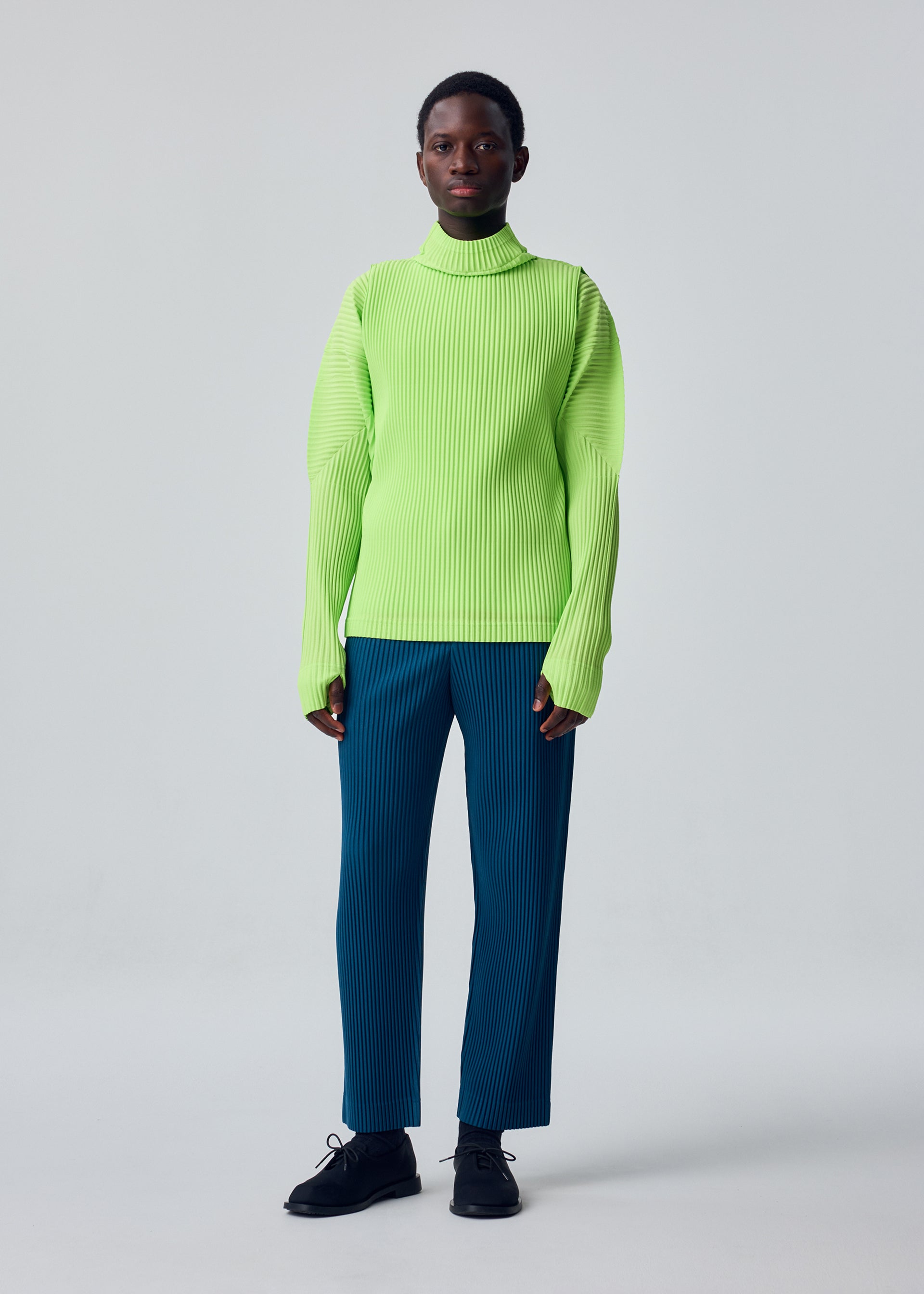 Zoomed view A person stands against a plain background wearing a bright green sporty long-sleeved ribbed turtleneck sweater with thumbhole sleeves, blue COLOR PLEATS 1 trousers by HOMME PLISSÉ ISSEY MIYAKE, and black shoes.