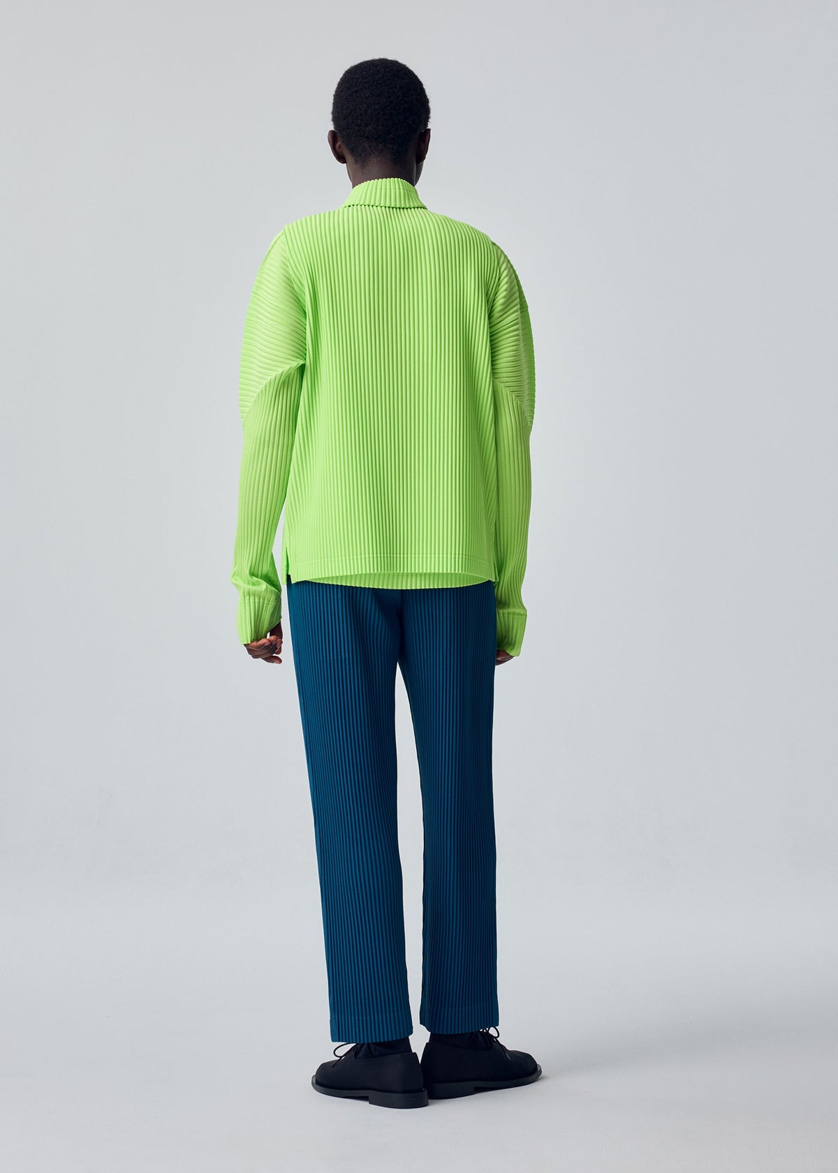A person stands facing away, wearing the HOMME PLISSÉ ISSEY MIYAKE COLOR PLEATS 1 blue pleated pants paired with a bright green ribbed long-sleeve sporty top and black shoes, set against a plain light gray background. view 7 of 8